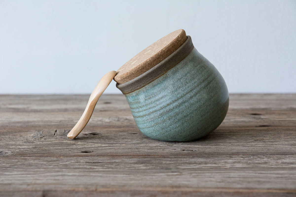 A small handmade ceramic jar with a light sage green glaze, a natural wood lid, and a matching wooden spoon resting on a rustic wooden table against a light gray background.