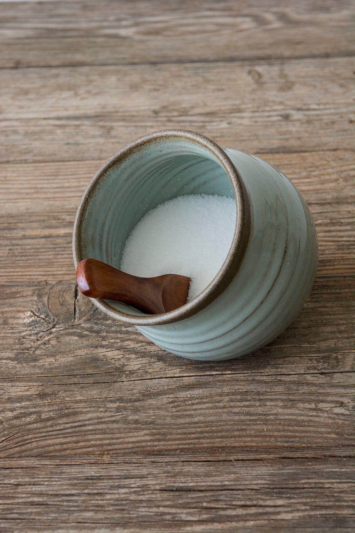 A light blue-green ceramic salt jar filled with white salt, featuring a small dark brown wooden spoon resting inside, set on a rustic wooden surface.