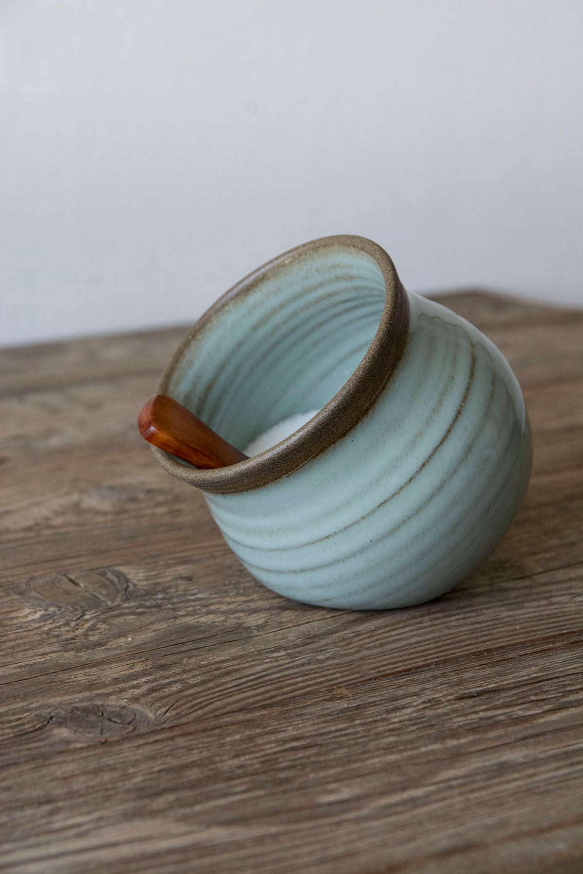 A small handmade ceramic cup with a light blue-green glaze and a brown rim, shown on a rustic wooden surface against a plain light background.
