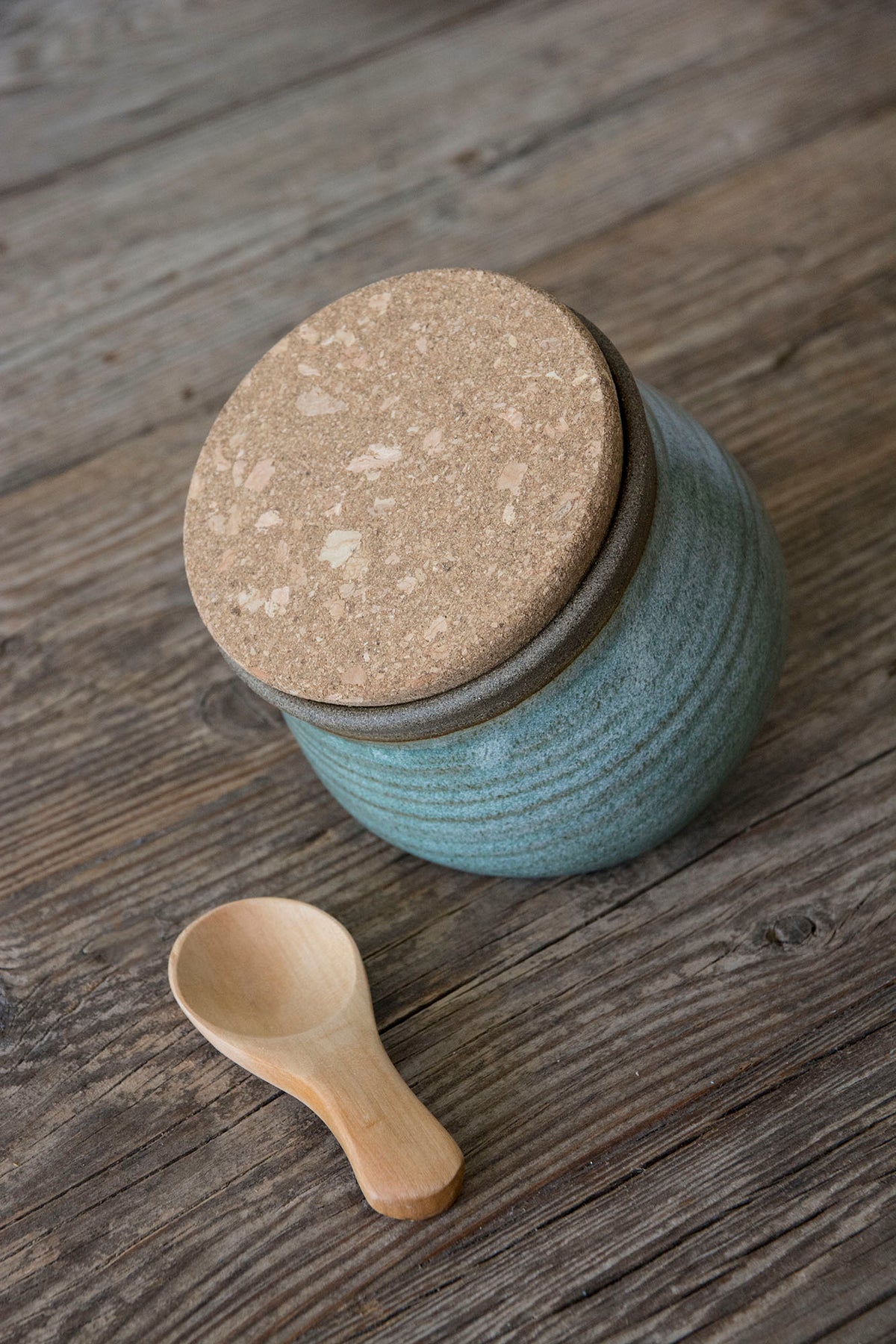 A handmade ceramic jar with a textured green sage matte body and a cork lid, resting on a rustic wooden surface next to a small wooden spoon.