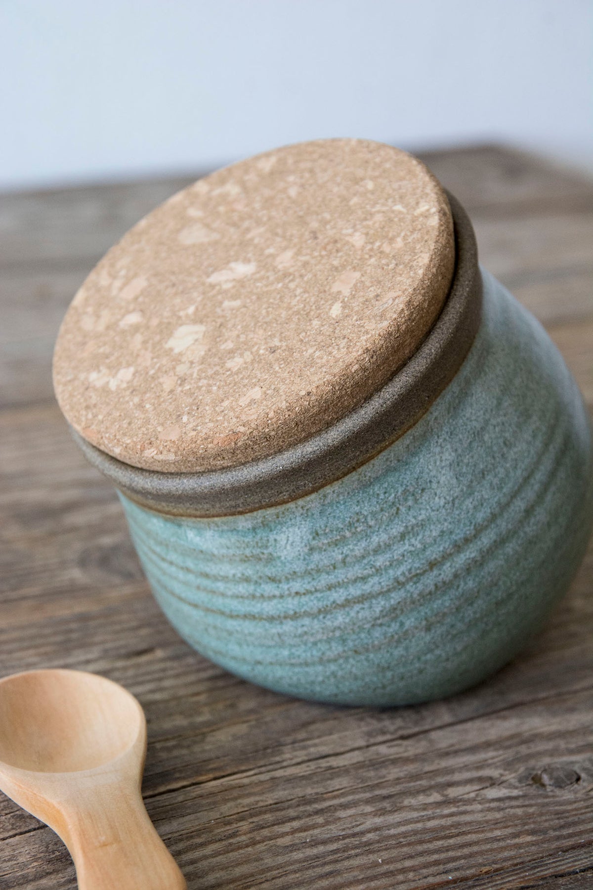 A handmade ceramic storage jar with a textured light blue-green glazed body, a brown ceramic band, and a round cork lid, shown on a rustic wooden tabletop with a wooden spoon in the foreground.