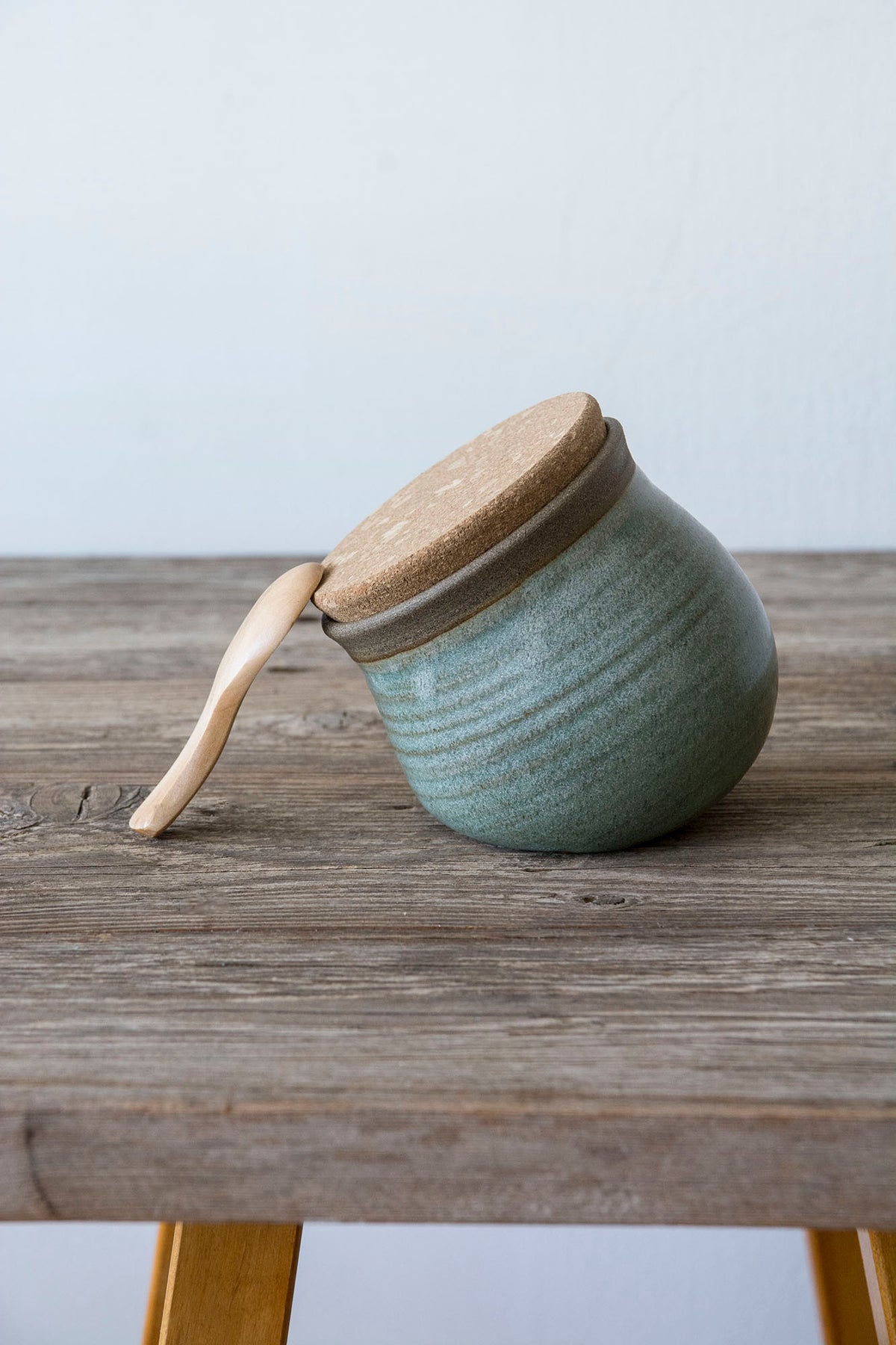 A small round ceramic jar with a light wood lid and a matching wooden spoon handle, resting on a rustic wooden table against a pale wall.