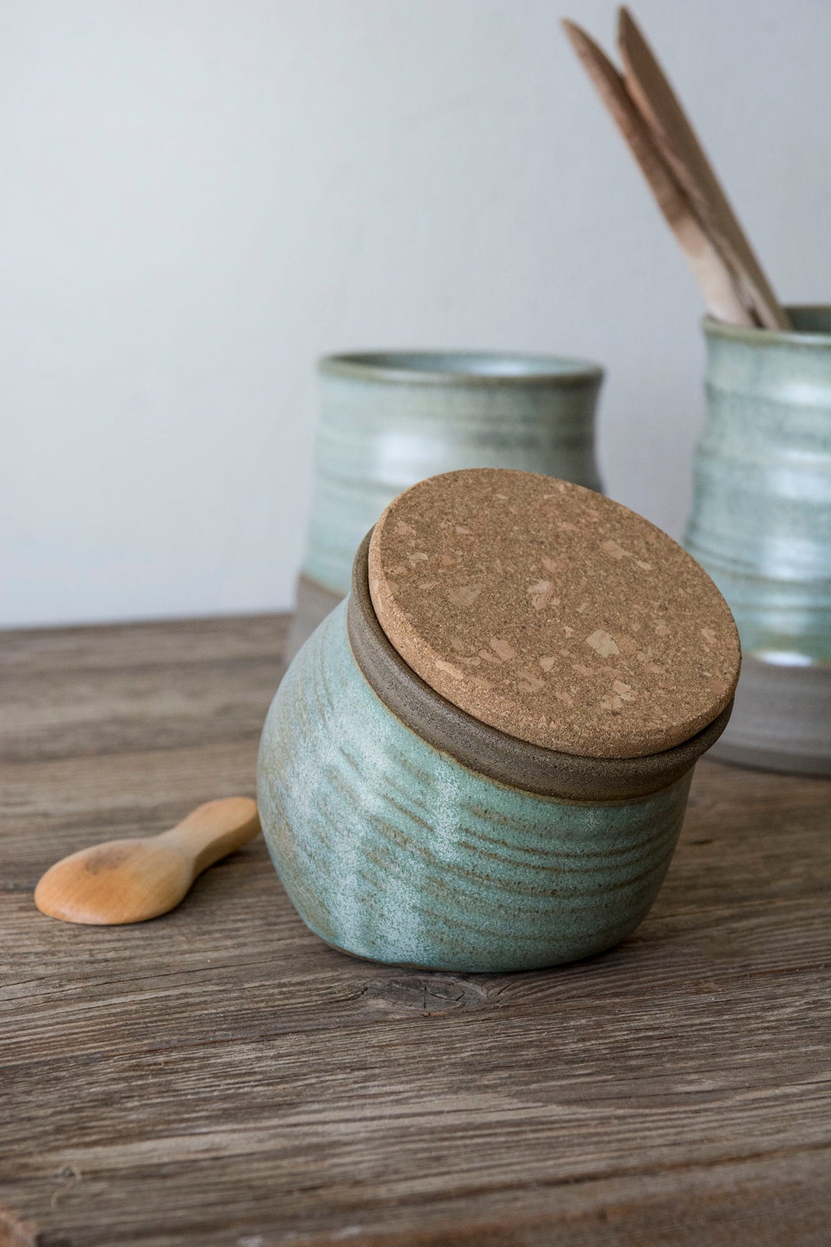 A handmade ceramic spice jar with a textured sage green glaze and a cork lid, sitting on a rustic wooden table next to a small wooden spoon. In the background, other matching ceramic containers are visible.